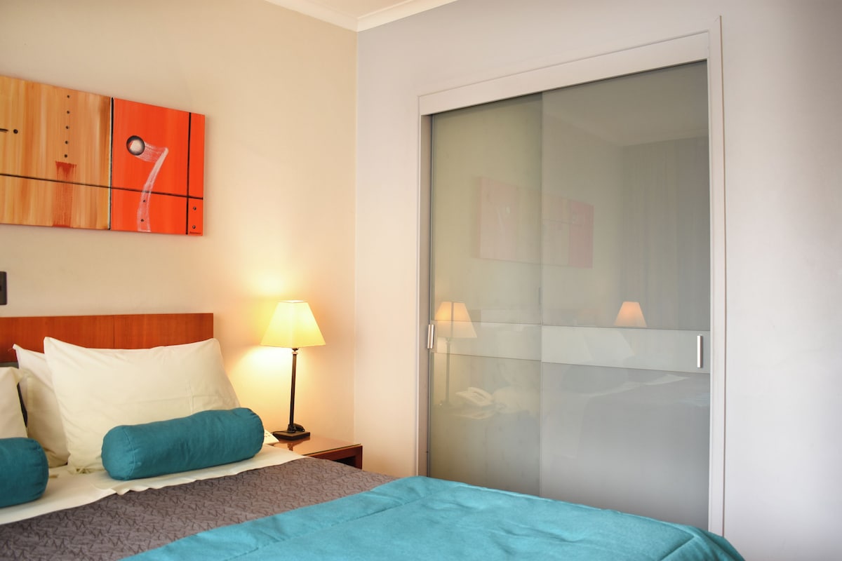 A hotel room with a bed, a nightstand with a lamp, a wardrobe with a sliding door, and a wall with a painting.