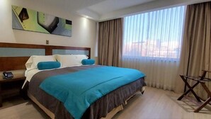 Ein Hotelzimmer mit einem großen Bett, einem Nachttisch, einem Sessel und einem Fenster mit Vorhängen.