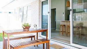 A patio with a wooden table and benches, a potted plant, and a sliding glass door leading to a kitchen area with stools.