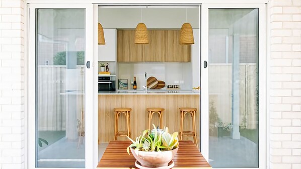 Una cocina moderna con armarios de madera, un comedor con mesa y sillas, y un ventanal corredero que da a un patio.