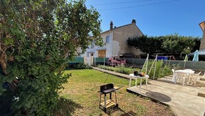 A backyard with a grassy lawn, a black grill, a white table with chairs, and a house in the background. - A pleasant room with shower room, 160cm bed and all comforts included. (Lavelanet)