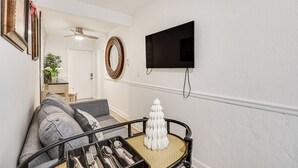 A living room with a sofa, a small table with cutlery, a decorative tree, a wall-mounted TV, and a ceiling fan. - Hollywood Hideaway: Close to Dtwn & Beaches (Hollywood)
