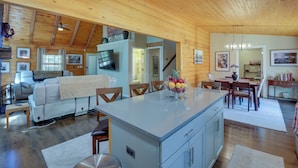 Ein geräumiger Wohnbereich mit Holzbalkendecke, einem Esstisch mit Stühlen, einer Kücheninsel mit Obstschale und einer Couch.