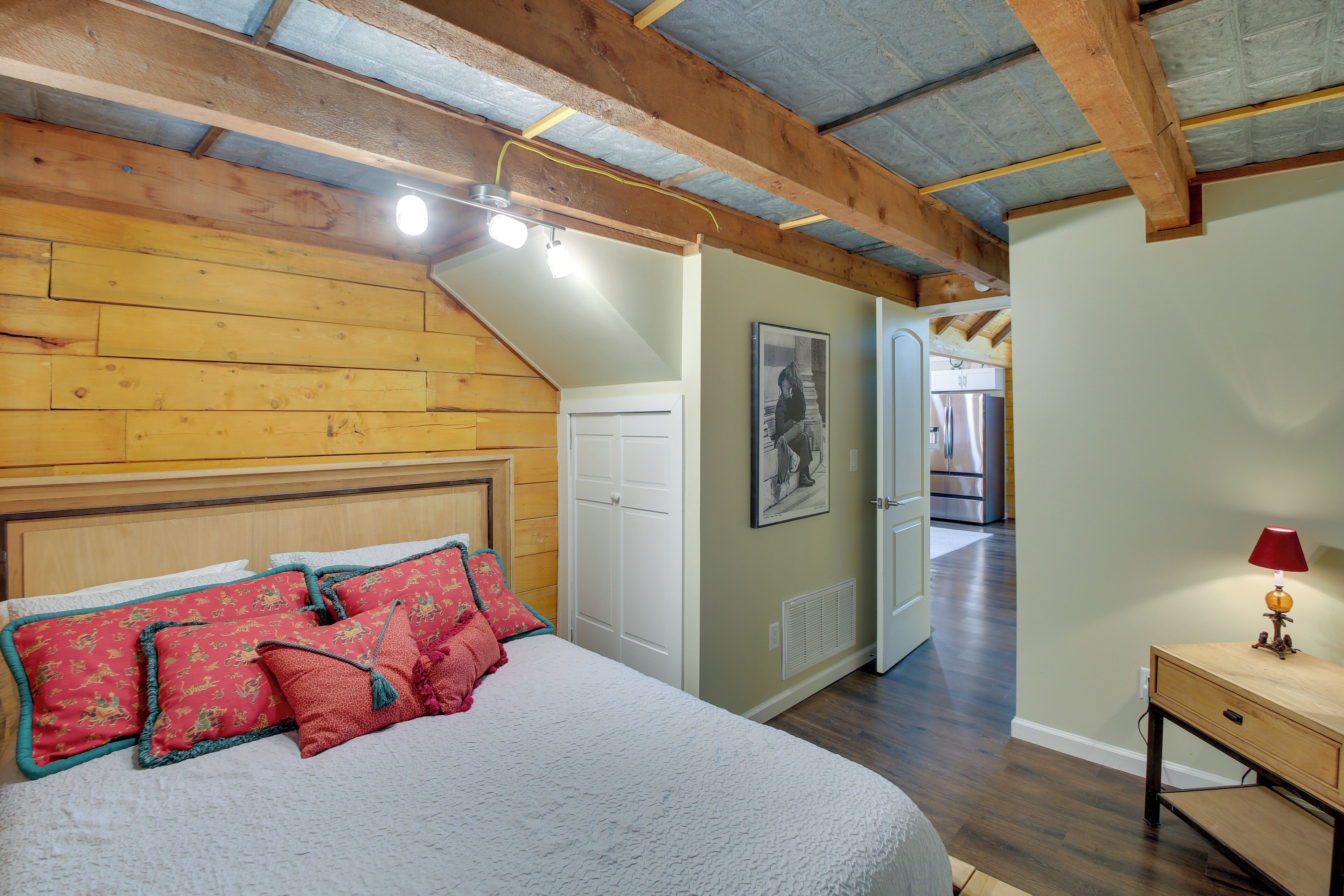 Une chambre avec un plafond en bois, un lit avec des oreillers rouges et verts, une porte blanche et un couloir menant à la cuisine.