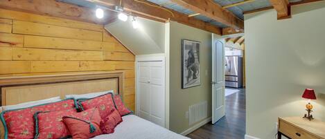 Une chambre avec un plafond en bois, un lit avec des oreillers rouges et verts, une porte blanche et un couloir menant à la cuisine.