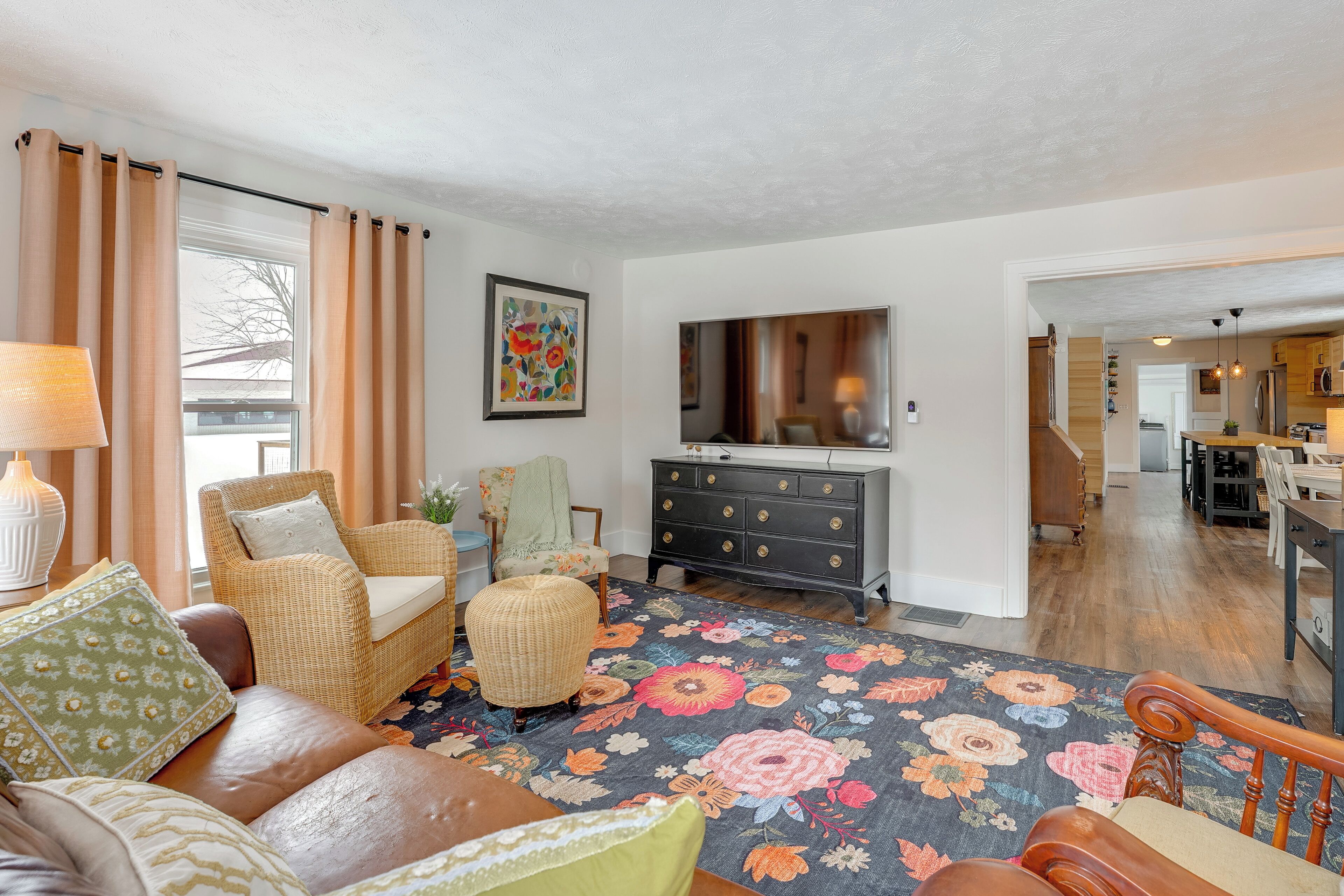 A living room with a sofa, armchair, ottoman, and a floral rug. There is a television on the wall and a wooden cabinet.