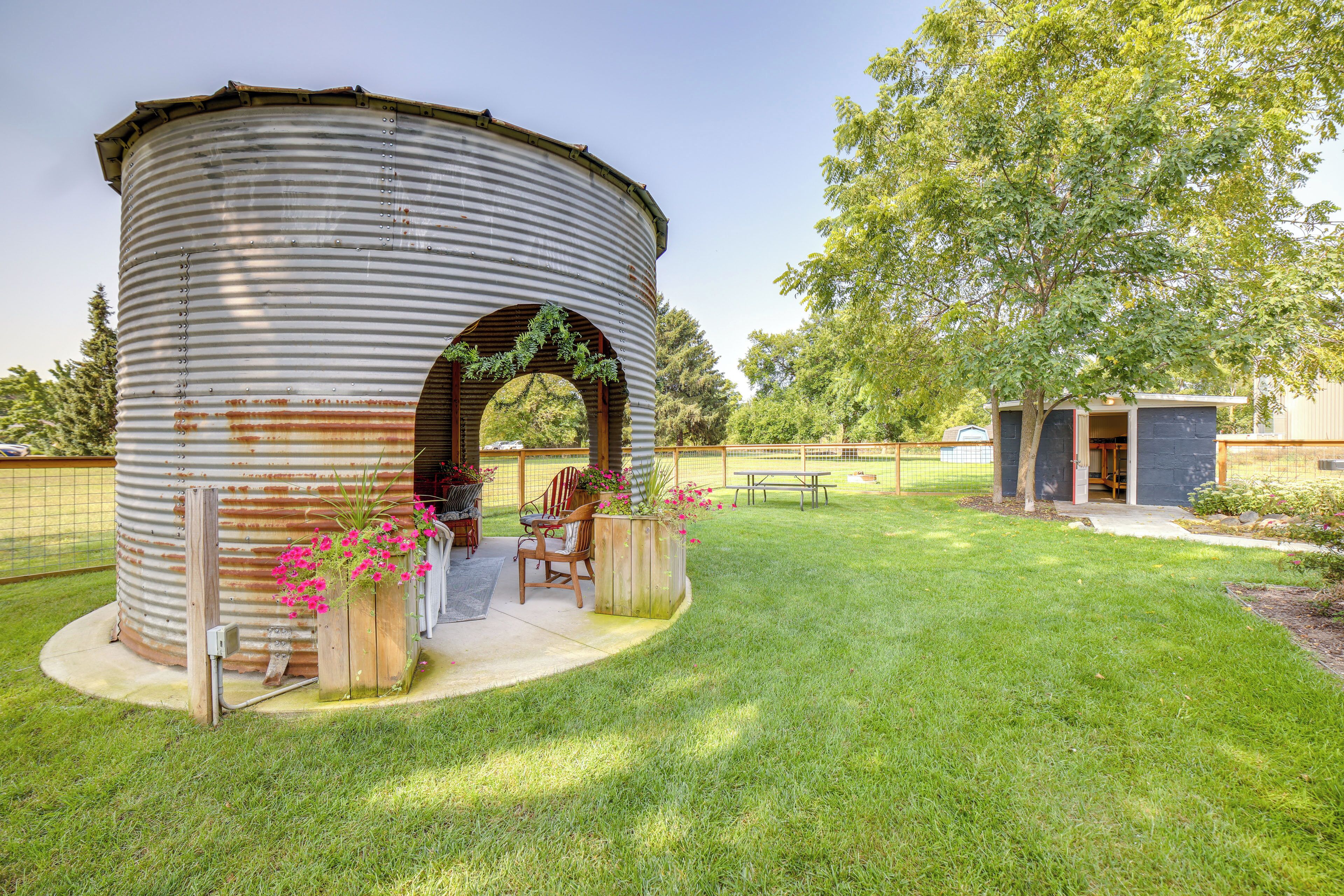 A rustic metal structure with a wooden bench and chairs, surrounded by a well-maintained lawn and a tree.