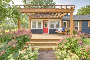 Un porche de madera con una puerta roja, rodeado de plantas en flor y una pérgola con luces colgantes.
