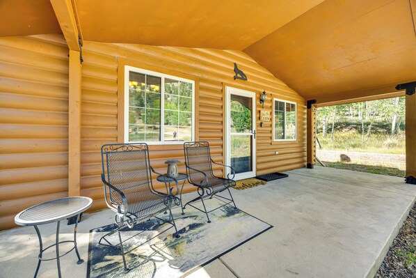 A covered porch with two metal chairs and a small table, wooden log cabin walls, and a door leading to a cabin. - Cabin in Eleven Mile Village w/ Patio & Gas Grill! (Lake George)