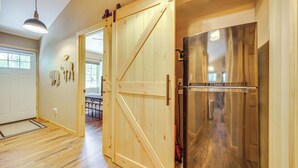 A spacious room with wooden flooring, a large wooden door, a stainless steel refrigerator, and a hanging light fixture.