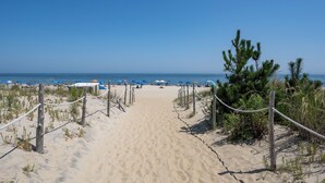 En sti langs en sandstrand, der fører til strandparasoller under en klar, blå himmel.