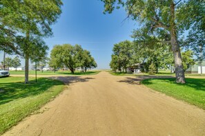 A dirt road flanked by trees and buildings, leading towards a body of water. - Yale Home in the Heart of Pheasant Hunting Country (Yale)