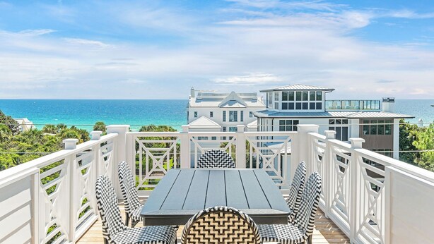 A deck with a table and chairs overlooking a large ocean view, a modern building with a glass facade, and a white railing.
