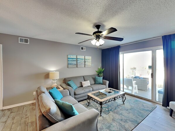 A living room with a grey sofa, a coffee table, and a ceiling fan. There are two windows with blue curtains, and a plant on the windowsill.