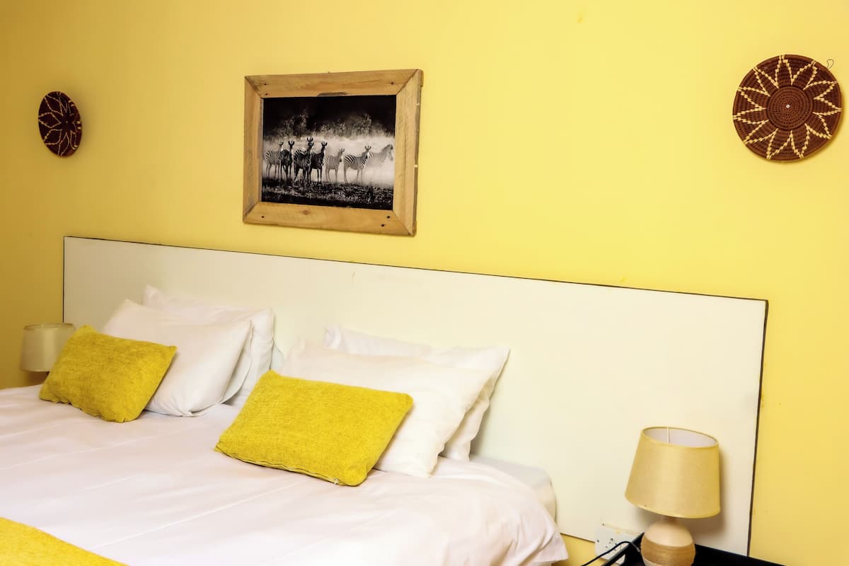A bed with white and yellow pillows, a wooden headboard, a wall-mounted picture, and a bedside lamp.