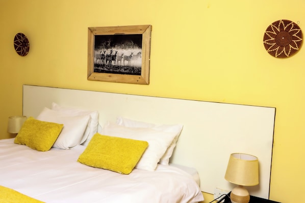 A bed with white and yellow pillows, a wooden headboard, a wall-mounted picture, and a bedside lamp. - Destiny Blo Inn (Kachikau)