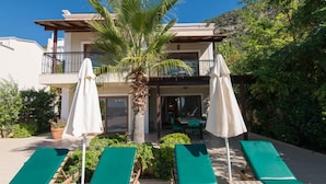 Un edificio de dos pisos con balcón, zona de estar exterior con sillones verdes y sombrillas, y una palmera.