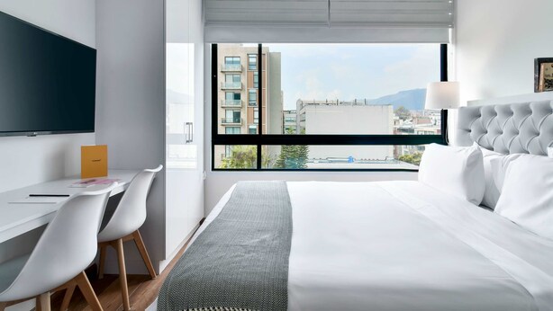 A modern hotel room with a large bed, a desk with chairs, a flat-screen TV, and a view of the city through large windows. - w Stylish Loft w Brilliant Deco in Chapinero (Bogotá)