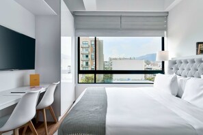 A modern hotel room with a large bed, a desk with chairs, a flat-screen TV, and a view of the city through large windows.