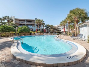 A swimming pool with a 5 ft 9 in depth, surrounded by lounge chairs and palm trees, with a building in the background. - Seaside Haven Stylish Condo 3 Pools New Rental (St. Augustine)