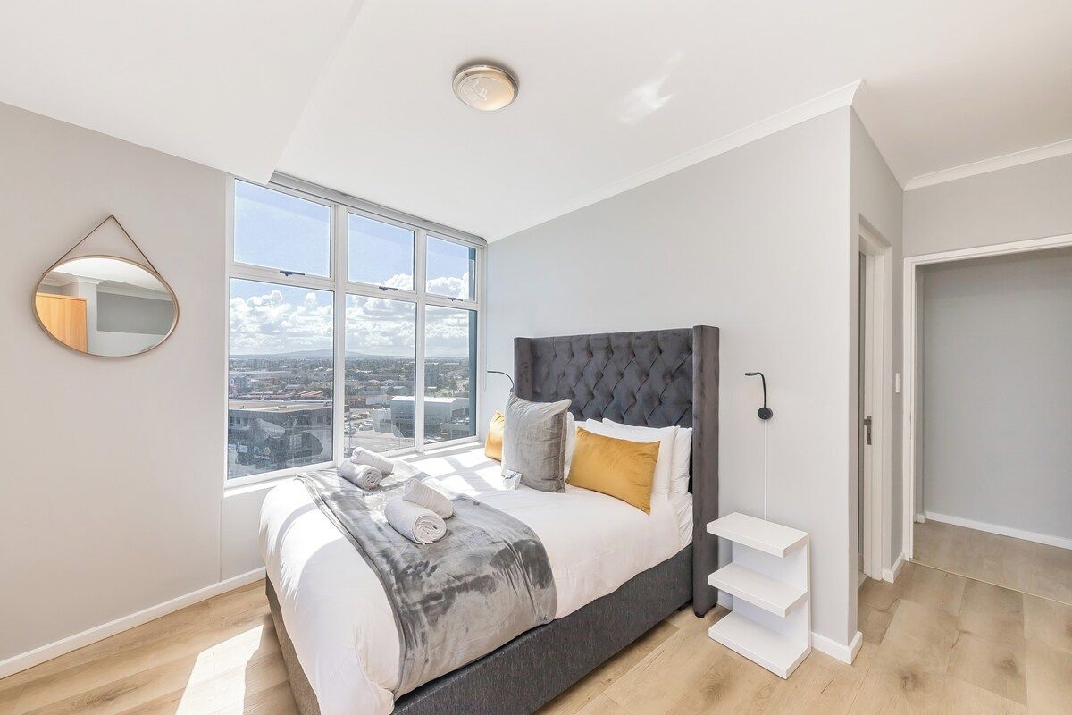 A bedroom with a large window, a bed with a grey headboard, and a white bedside table.