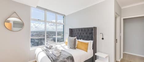 A bedroom with a large window, a bed with a grey headboard, and a white bedside table.