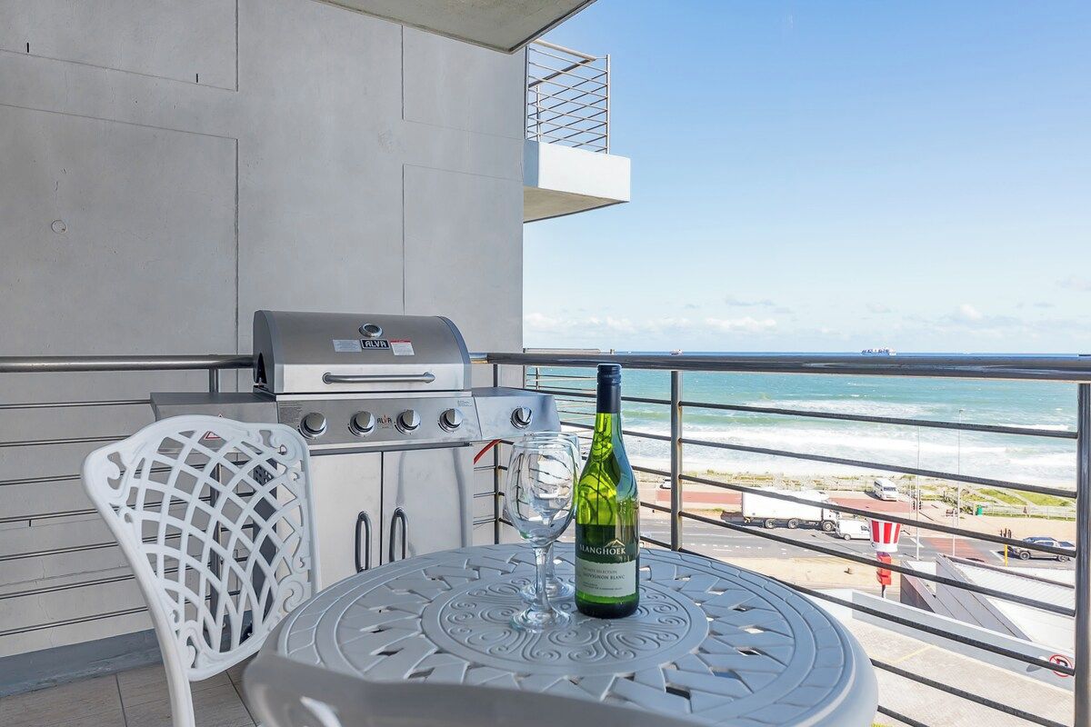 Balkon med grill, vinflaske og glas på et bord, med udsigt over strand og hav.
