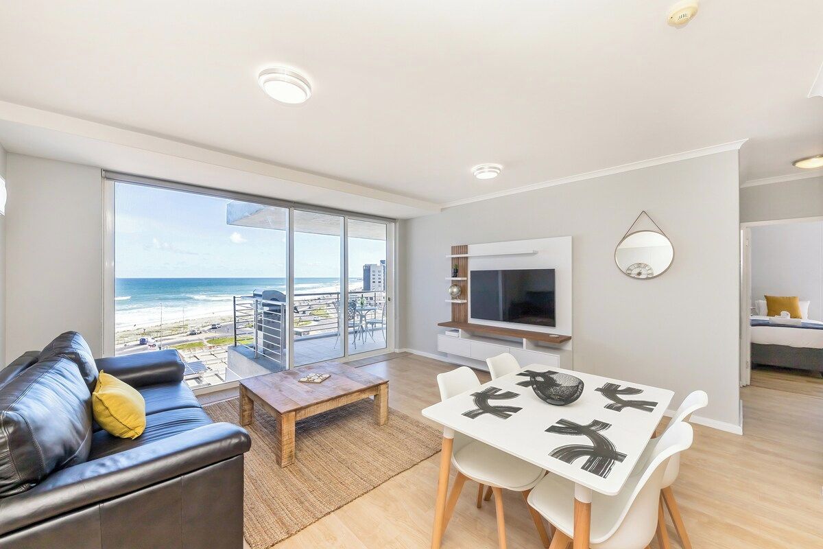 Una sala moderna con un gran ventanal con vistas a la playa, un televisor de pantalla plana y un comedor con una mesa y sillas blancas.