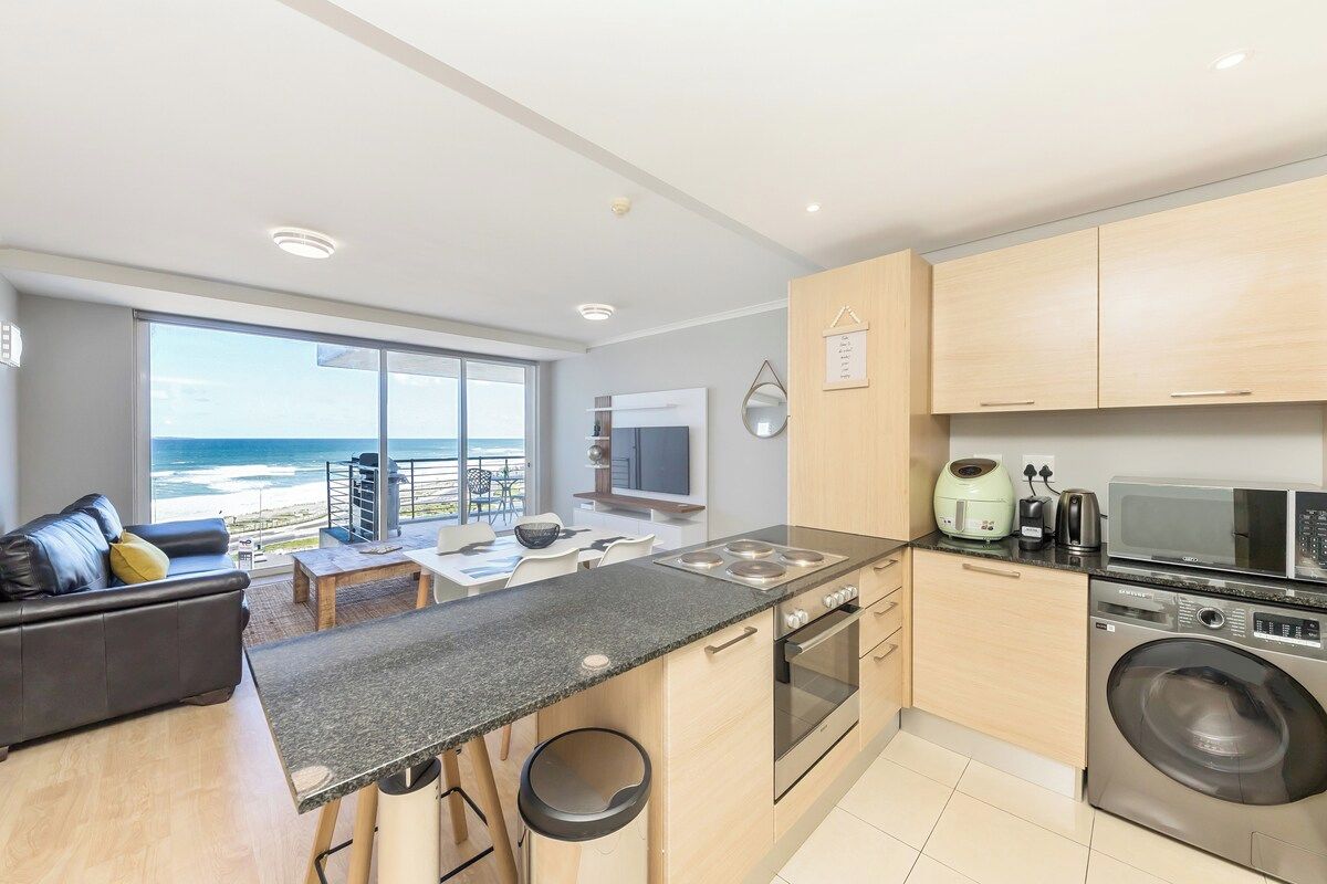 A modern kitchen with a large island, a washing machine, and a view of the ocean.