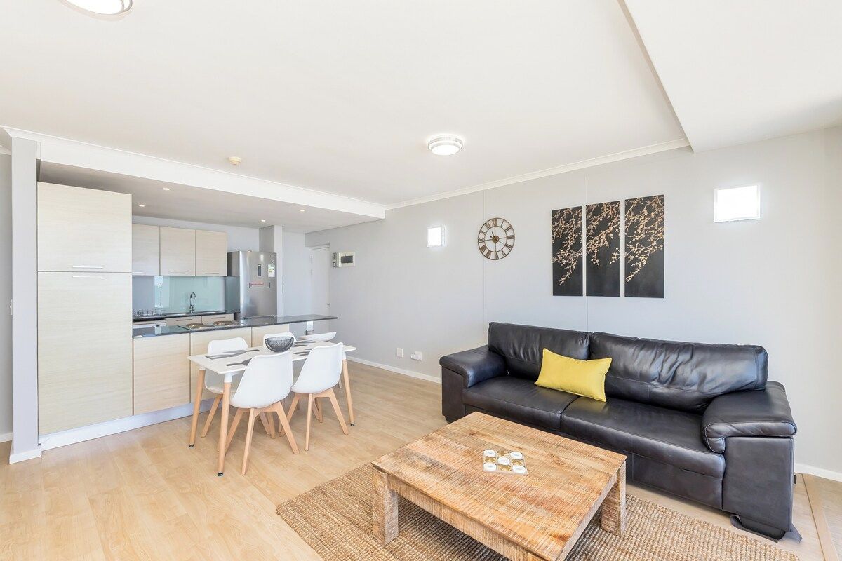 A modern living room with a black leather sofa, a wooden coffee table, and a dining area with white chairs.
