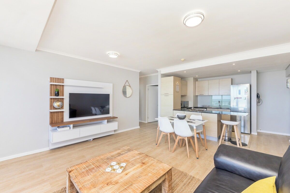 A modern living room with a flat-screen TV, a dining area with chairs, a sofa, and a kitchen in the background.