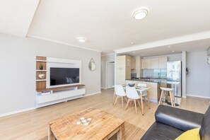 A modern living room with a flat-screen TV, a dining area with chairs, a sofa, and a kitchen in the background.