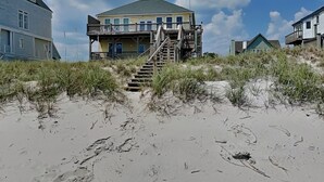 Una casa sulla spiaggia, con una scala che conduce a un balcone, immersa nella sabbia e nella vegetazione.