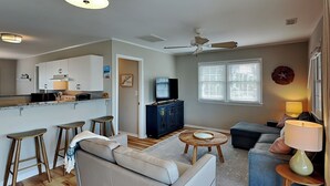 A modern living room with a sofa, coffee table, and a television. There is a kitchen area with white cabinets and a bar counter. - The Sassy Sea Turtle | Spacious + Pet-Friendly Retreat, Just Steps From The Beac (Topsail Beach)