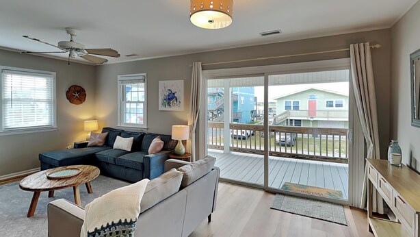 A spacious living room with a grey sofa, a coffee table, and a ceiling fan. A large window offers a view of a deck and buildings outside. - The Sassy Sea Turtle | Spacious + Pet-Friendly Retreat, Just Steps From The Beac (Topsail Beach)