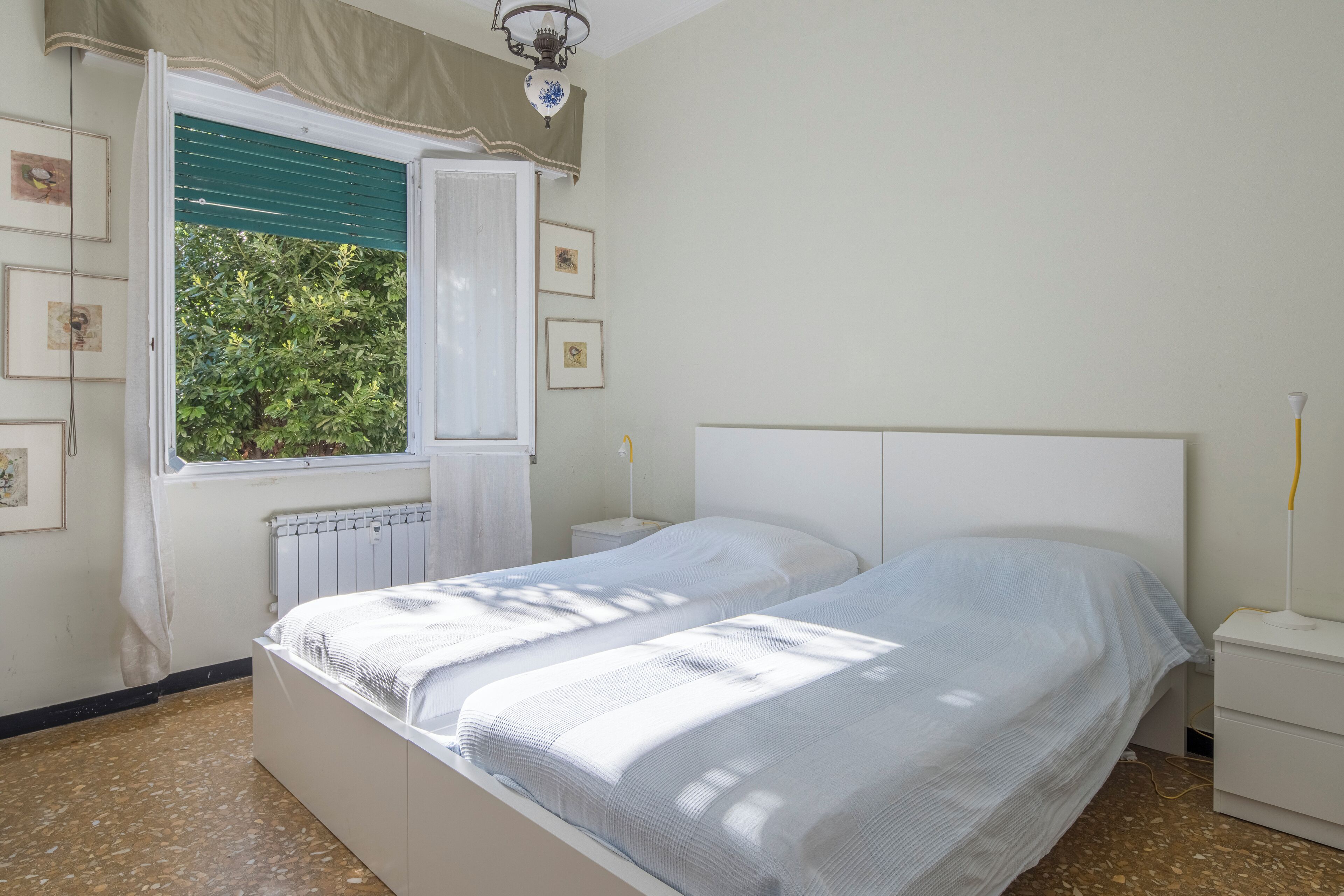 A bedroom with two beds, a window with curtains, a radiator, and framed pictures on the wall.