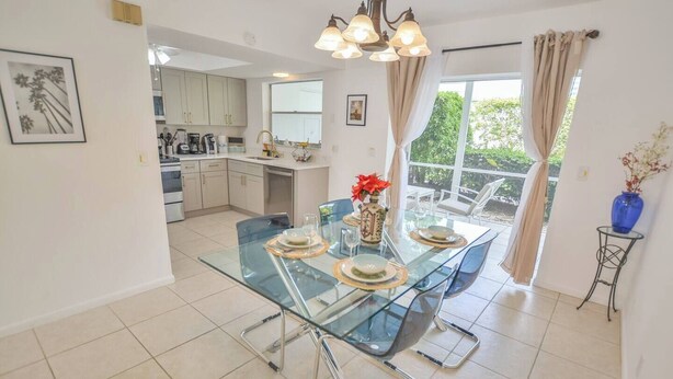 A modern kitchen with white cabinets, a dining area with a glass table and chairs, and a view of greenery outside. - Walk to the beach 2BR 2BA 1300SF (Riviera Beach)