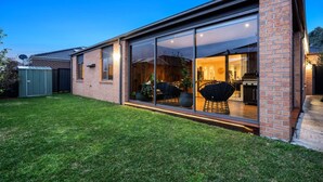 A modern house with a large glass sliding door leading to a patio area with outdoor furniture. - Modern Cozy Retreat, Park, Mall! (Berwick)
