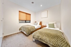 A bedroom with two single beds, a window with blinds, a bedside table, and a nightstand. - Spacious 4BR Family Retreat | Theatre & Sunroom (Berwick)