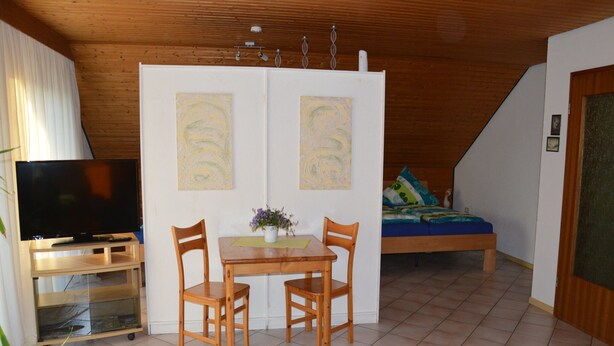 A room with a wooden ceiling, a TV on a stand, a dining table with chairs, a bed with pillows, and two abstract paintings on the wall. - DIANA vacation apartment 5 minutes from Europa Park (Rust)