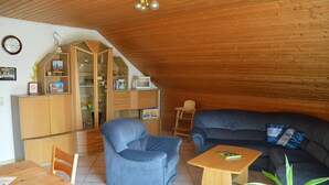 A cozy living room with a wooden ceiling, a blue armchair, a dark sofa, and a wooden coffee table.