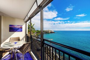 A balcony with wicker furniture, a glass table, and a painting. It overlooks a coastal area with a clear sky and calm sea.