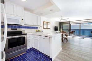 A modern kitchen with white cabinets, a built-in oven, and a dishwasher. There is a dining area with a table and chairs, and a view of the ocean through large windows.