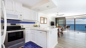 Una cocina moderna con gabinetes blancos, horno integrado y lavavajillas. Cuenta con un comedor con mesa y sillas, y vistas al océano a través de amplios ventanales.