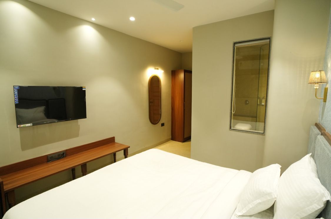 A hotel room with a bed, a TV mounted on the wall, a wooden bench, a mirror, and a shower area.