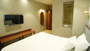 A hotel room with a bed, a TV mounted on the wall, a wooden bench, a mirror, and a shower area. - PACHMARHI TIGER  (Pipariya)