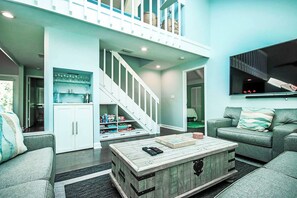 A spacious living room with a staircase leading to an upper level, a flat-screen TV mounted on the wall, and a coffee table with books and a remote control.