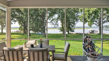 A covered patio with a table and chairs, large windows offering a view of a lake and trees.