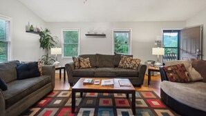 A living room with two sofas, a coffee table, and a patterned rug. There are two windows with blinds, a plant, and a shelf with decorative items. - Whispering Pines - Spectacular Views & Hot Tub 3 Bedroom Home (Burnsville)
