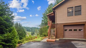 A two-story house with a wooden staircase leading to a balcony, a garage, and a driveway surrounded by greenery. - Whispering Pines - Spectacular Views & Hot Tub 3 Bedroom Home (Burnsville)